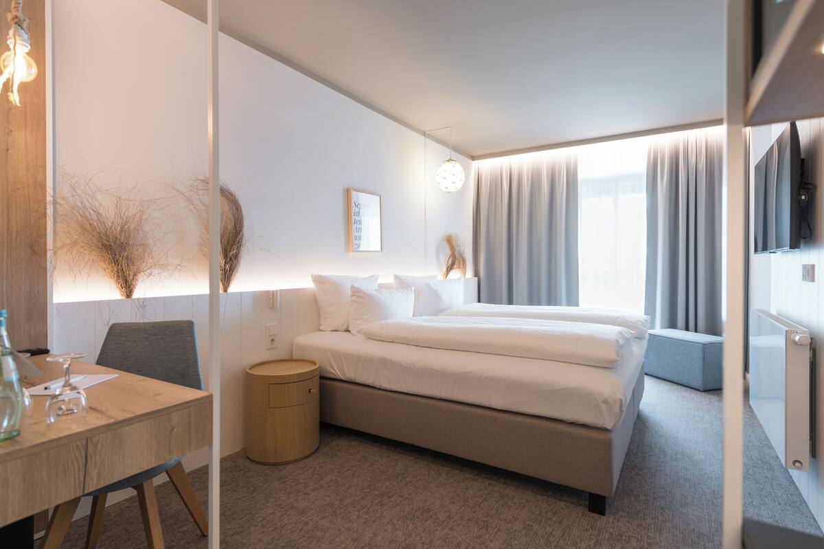 A modern hotel room with a bed, bedside table, lamp, and a view of the outdoors.