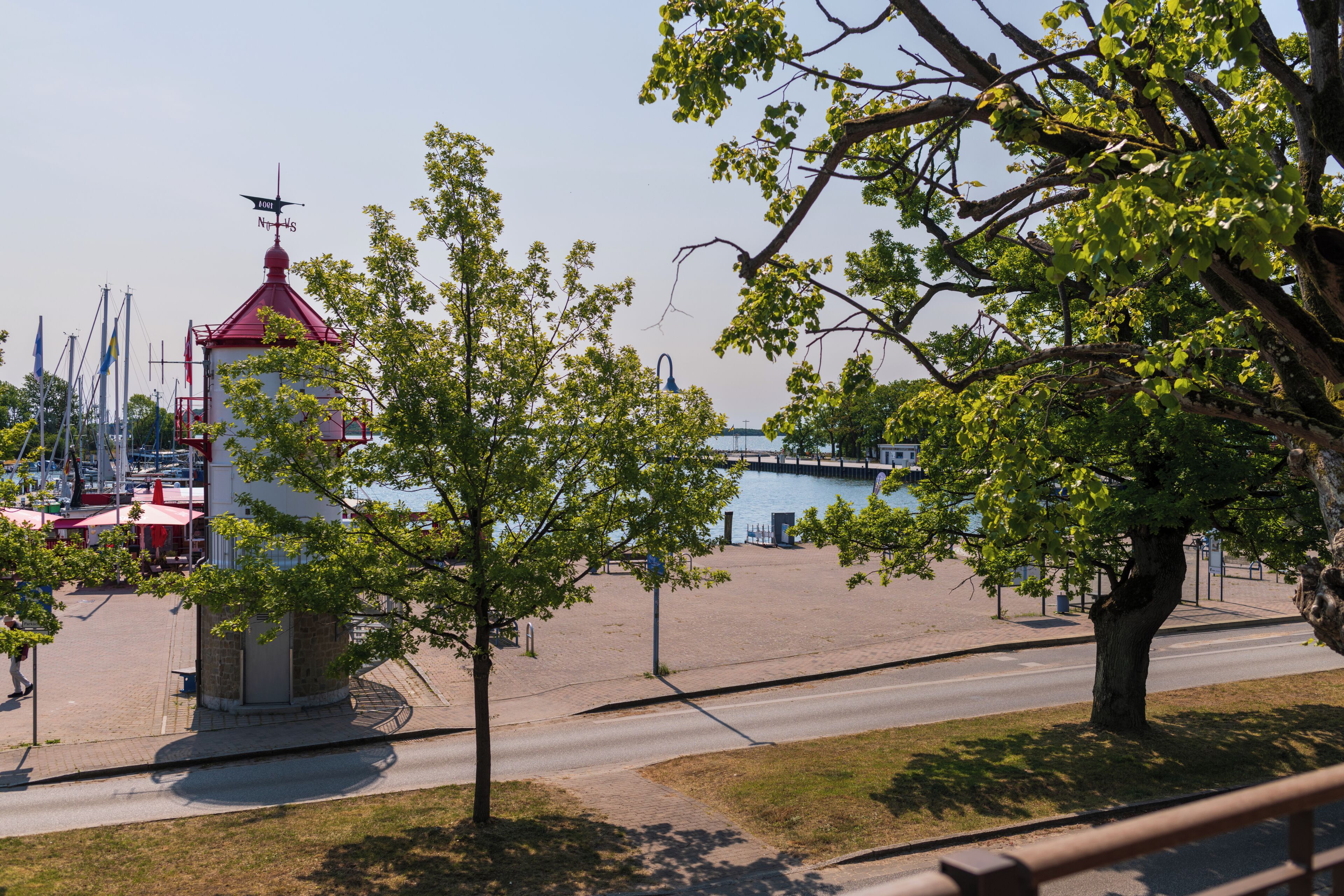 A view of a waterfront area with a red-roofed building, a tree-lined sidewalk, and a road.