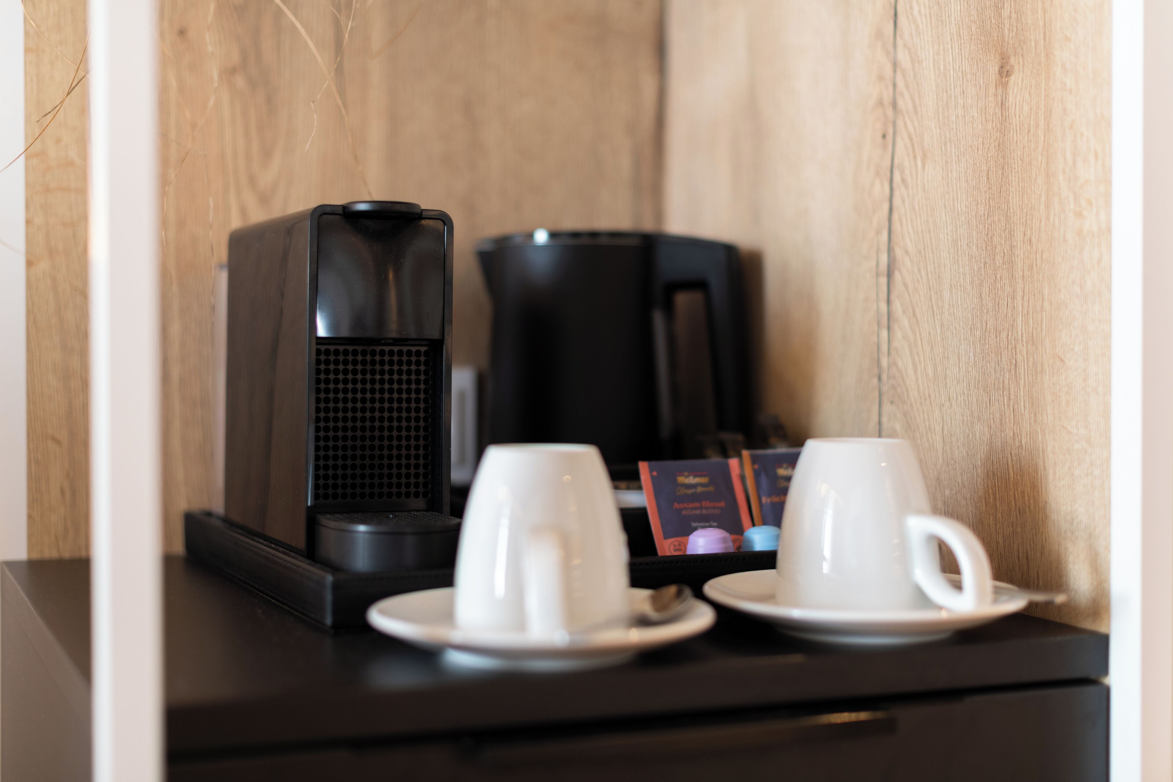 A coffee maker and two white cups on a black tray, with a wooden wall in the background.