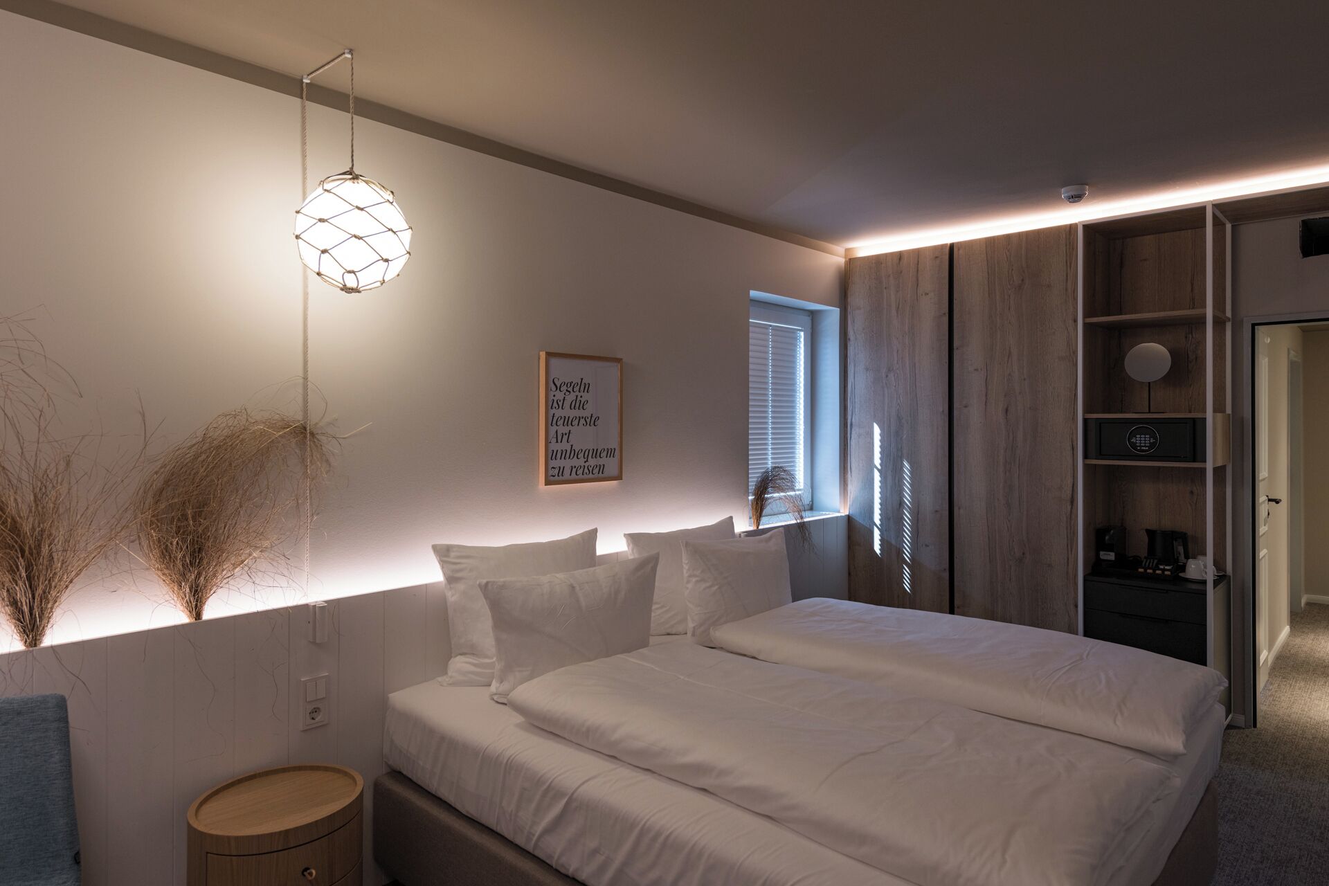 A bedroom with a bed, bedside table, wall-mounted light, window, and a framed quote.