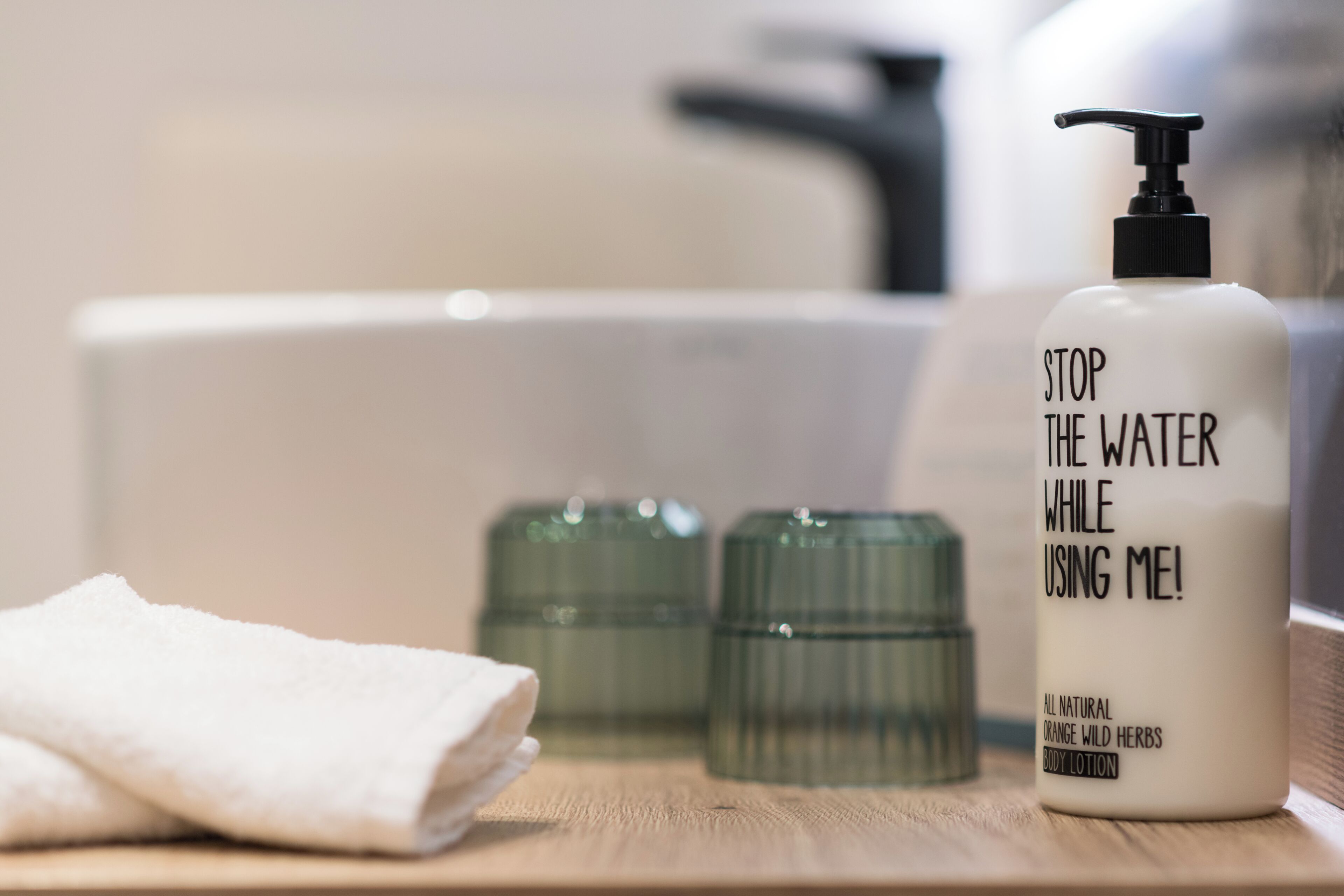 A bathroom with a white bottle labeled 'STOP THE WATER WHILE USING ME!', a white towel, and two green glass containers.