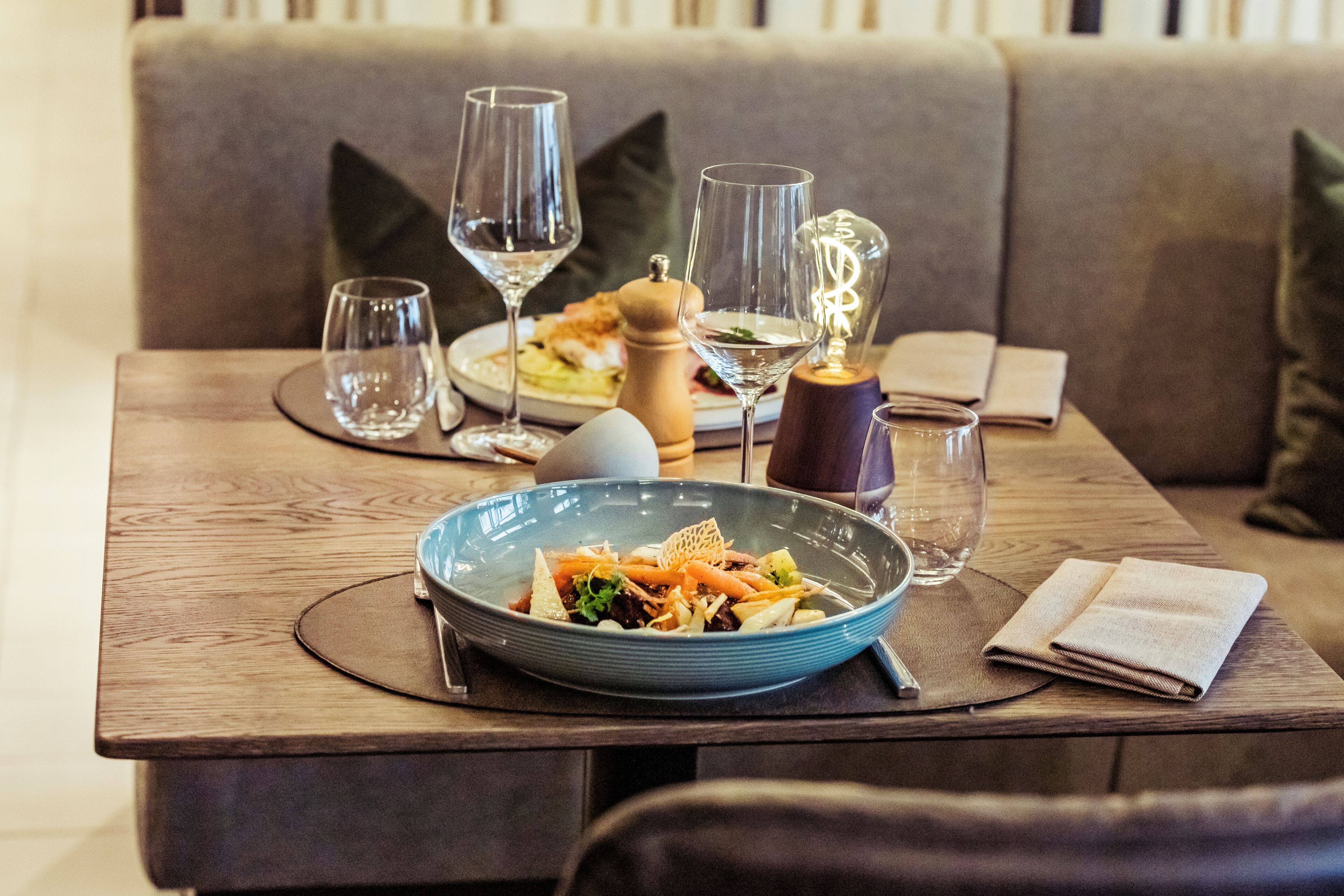 Een eettafel met een blauwe schaal sla, wijnglazen, een pepermolen en een servet.