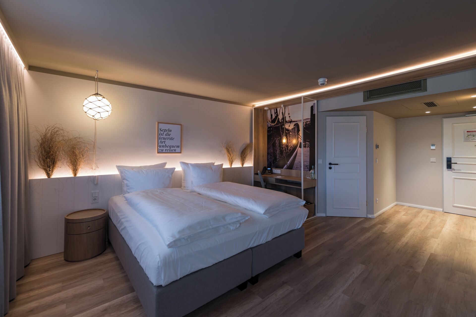 Un dormitorio moderno con una cama, mesitas de noche, una lámpara fijada a la pared y una puerta que da a otra habitación.