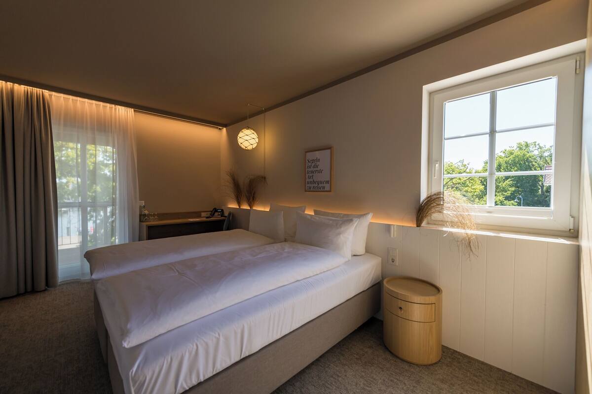 A bedroom with a bed, bedside table, lamp, window, and a small wooden stool.