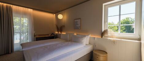 A bedroom with a bed, bedside table, lamp, window, and a small wooden stool.