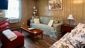 A room with a wooden paneled wall, a sofa with floral cushions, a red armchair, a wooden coffee table, a lamp, and a painting on the wall.