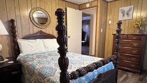 A bedroom with a wooden four-poster bed, a nightstand with a lamp, a dresser, a mirror, and a door leading to another room.