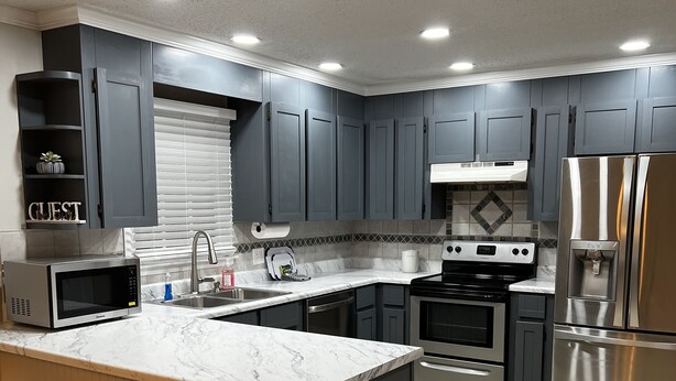 A modern kitchen with grey cabinets, a large island with bar stools, stainless steel appliances, and a tiled backsplash.
