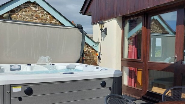 Et udendørs spabad med en baggrund af stenvæg, en træpergola og en glasdør, der fører til en terrasse med et rundt bord og stole.