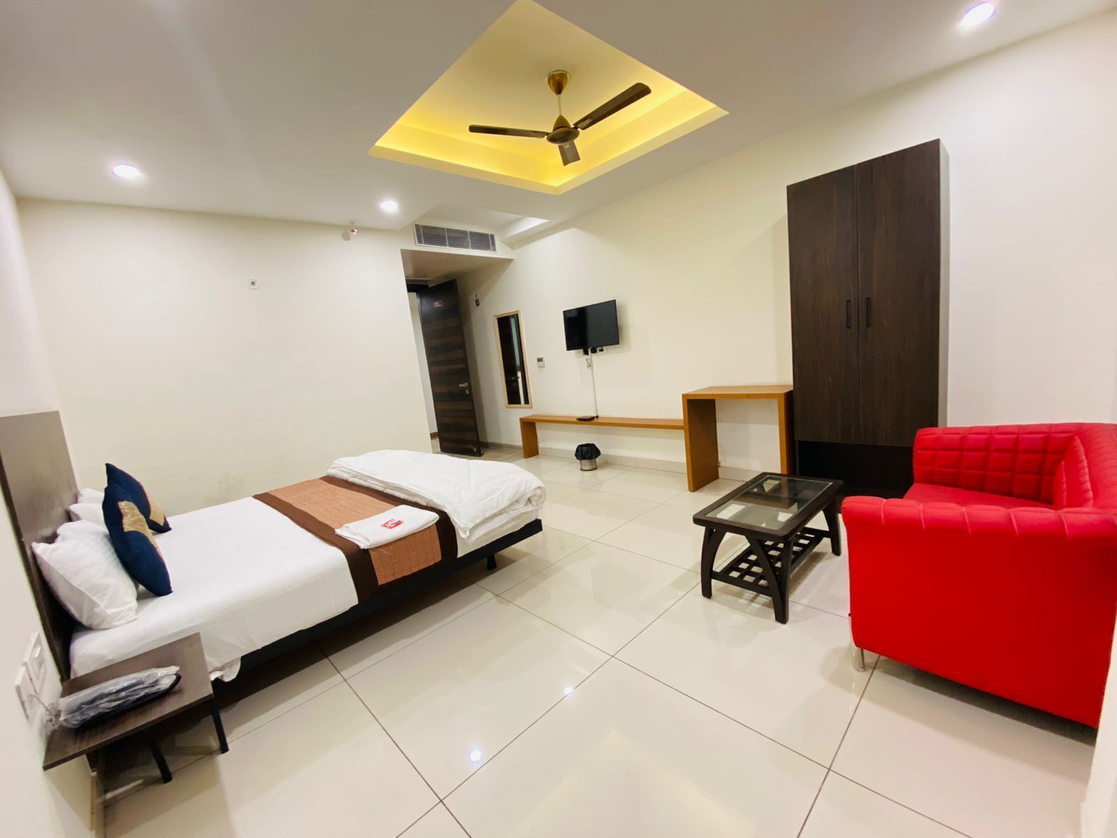 A modern hotel room with a bed, a red sofa, a small table, a ceiling fan, and a TV.