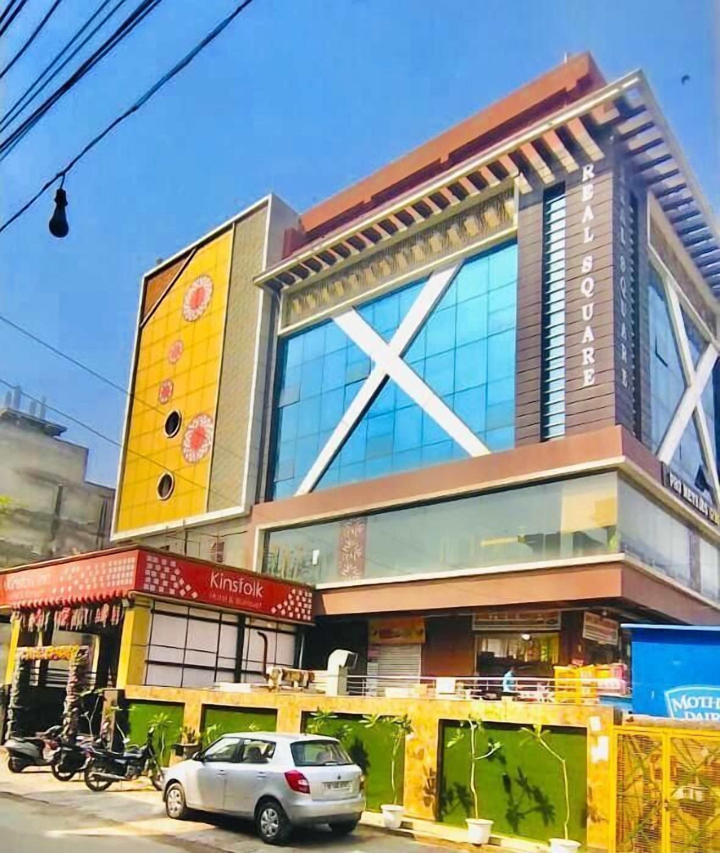 A modern building with a prominent 'REAL SQUARE' sign, a visible storefront named 'Kinsfolk', and a parked car in front.