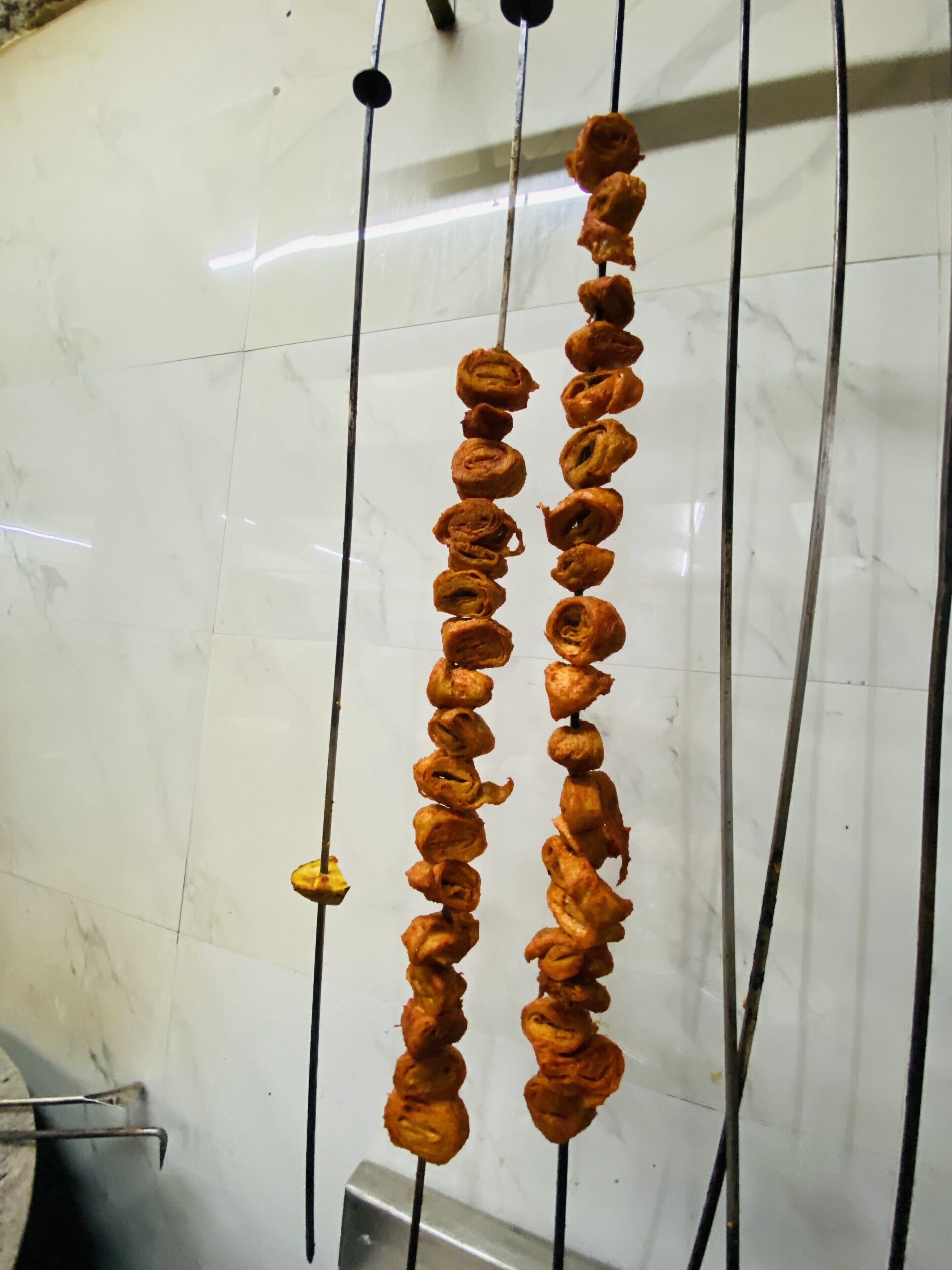 Una hilera de alimentos ensartados en una barra metálica, sobre un fondo de pared de azulejos blancos.