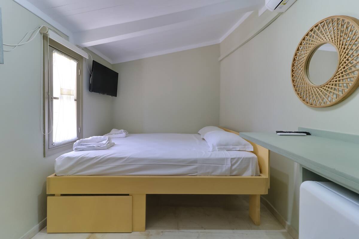 A small, neatly arranged bedroom with a bed, a bedside table, a TV mounted on the wall, and a window with curtains.