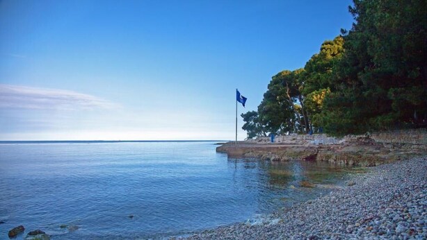 Une plage de galets avec une eau calme et un drapeau flottant sur un promontoire rocheux.