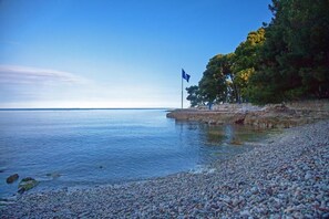 Una playa de piedrecitas con agua tranquila y una bandera en un promontorio rocoso.