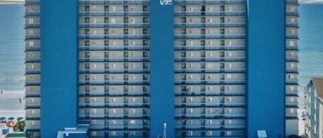 Un hotel grande con una torre central y dos alas, que cuenta con un estacionamiento con varios autos y palmeras en primer plano.