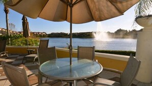 An outdoor patio with a round glass table and chairs, a large umbrella, and a view of a lake with a fountain. - 3000yl 4 Bedroom Townhouse by RedAwning (Kissimmee)