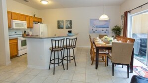 A compact kitchen with a central island, wooden cabinets, a microwave, and a refrigerator. There is a dining area with a table and chairs, and a large window providing a view. - 3000yl 4 Bedroom Townhouse by RedAwning (Kissimmee)