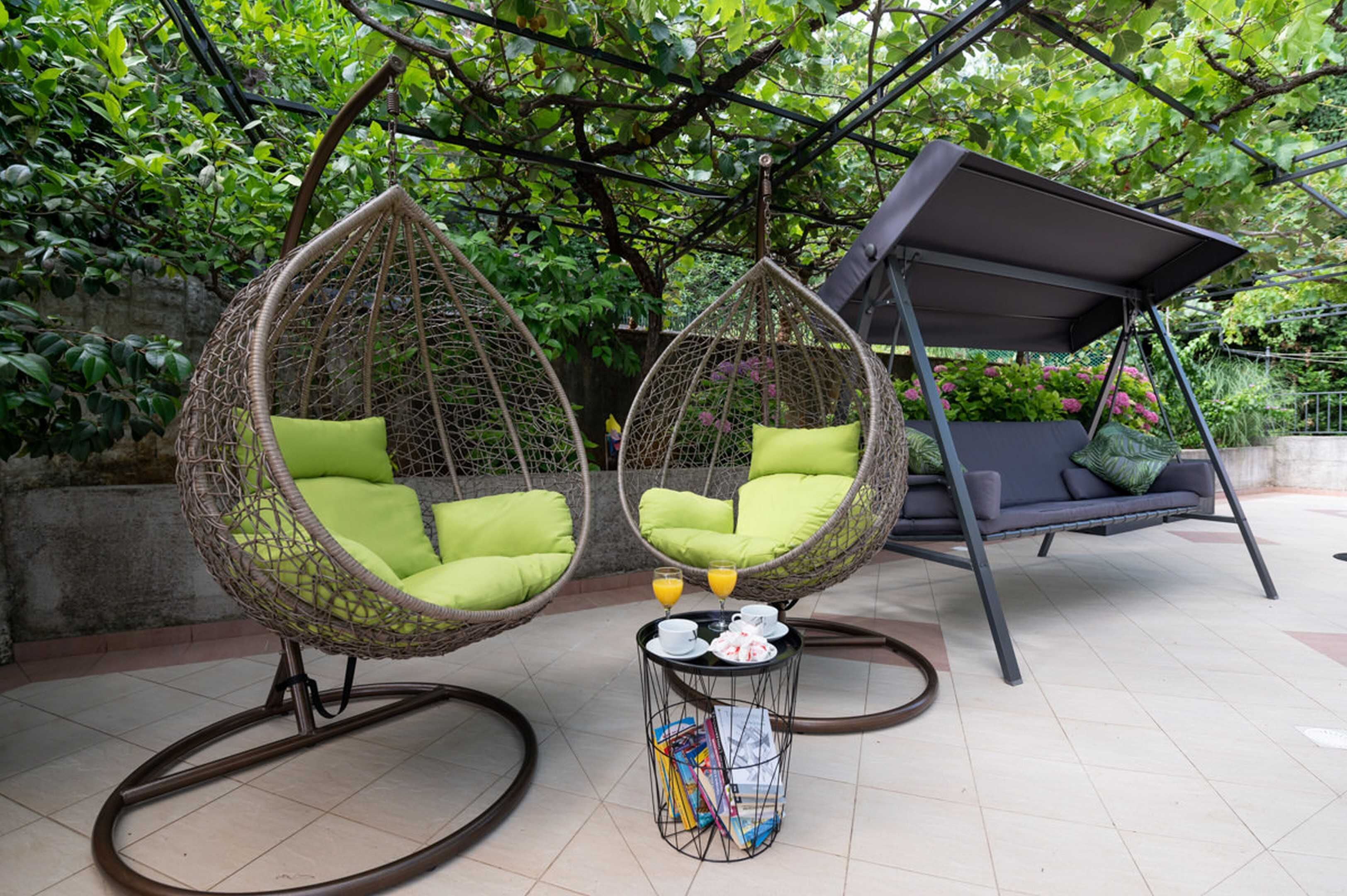 Two hanging chairs with green cushions, a small table with a drink and a magazine, and a swing chair in a garden setting.