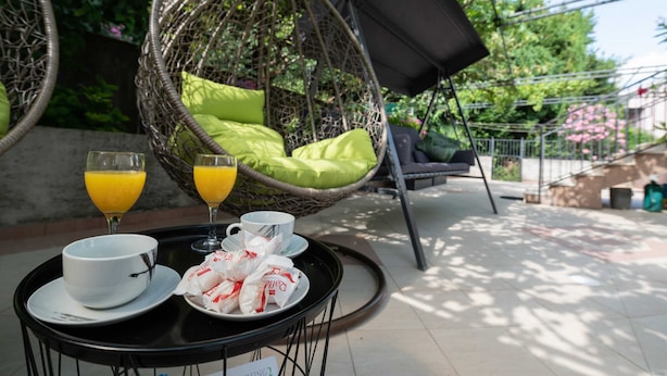 Una silla colgante de mimbre con cojines verdes, una mesa con una bandeja de alimentos para el desayuno, incluyendo un tazón, tazas y un vaso de jugo de naranja, con un fondo de vegetación y un edificio.