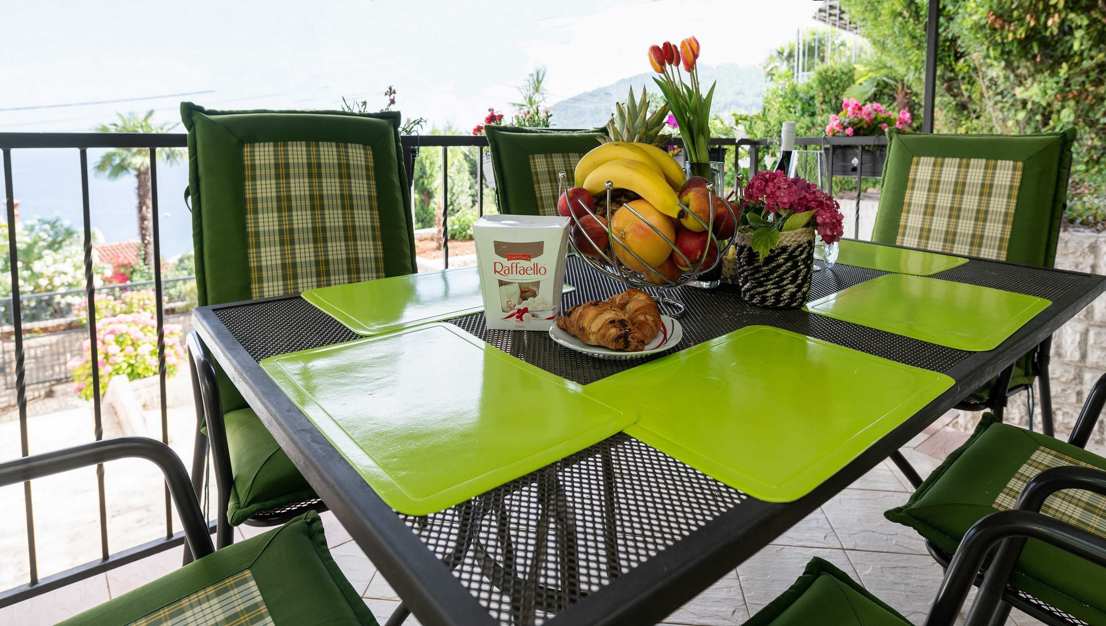 A table with a green placemat, a box of Raffaello coffee, a plate with a croissant, and a fruit bowl with bananas, apples, and oranges.