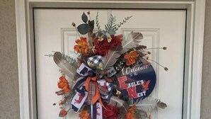 Eine Tür mit einem festlichen Kranz, der ein großes Schleifenband, ein Willkommensschild und verschiedene Blumen zeigt.
