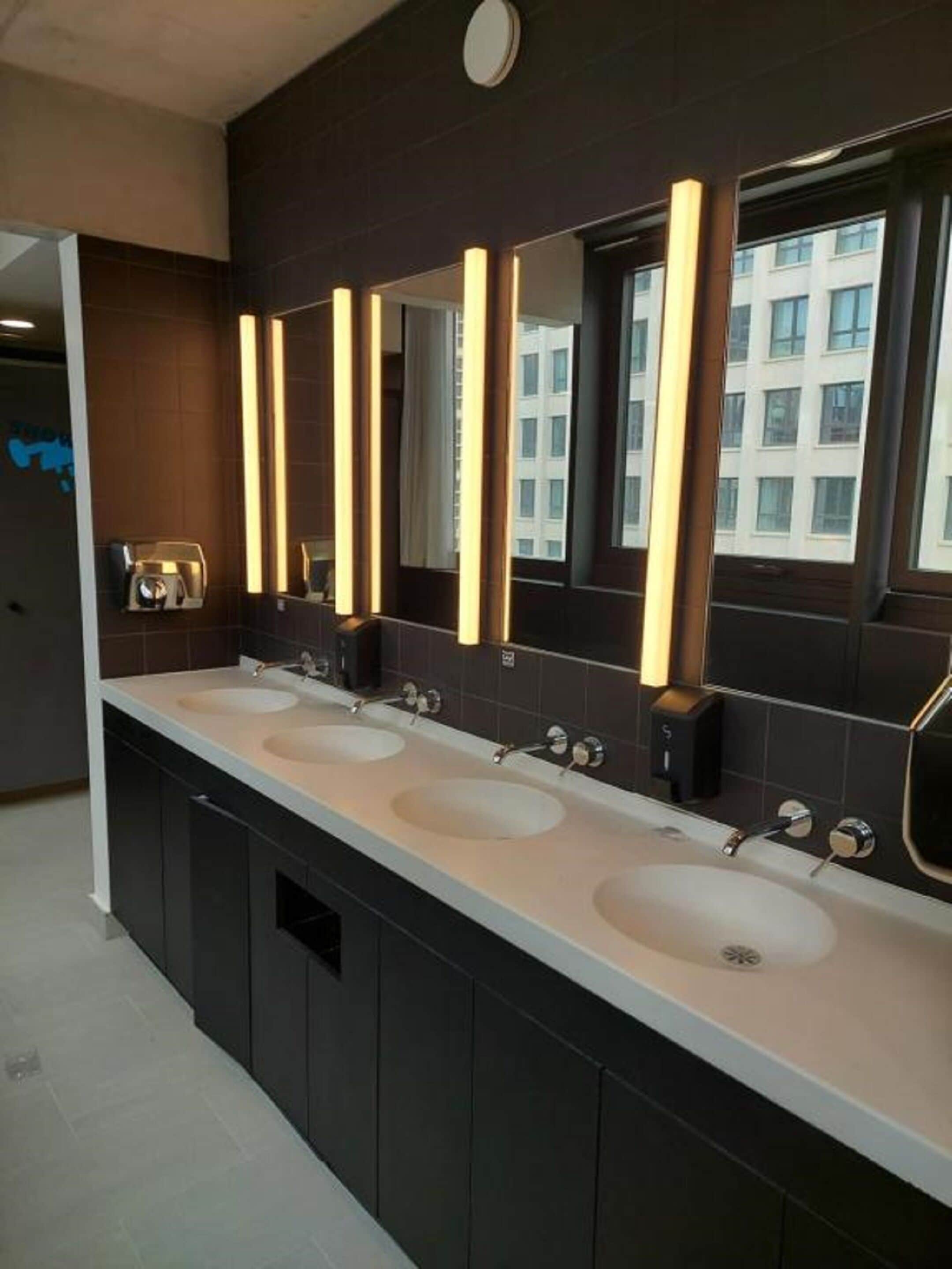 Une salle de bain moderne avec trois lavabos, des murs recouverts de carreaux foncés et de grands miroirs dotés de bords éclairés.