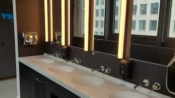 Une salle de bain moderne avec trois lavabos, des murs recouverts de carreaux foncés et de grands miroirs dotés de bords éclairés.