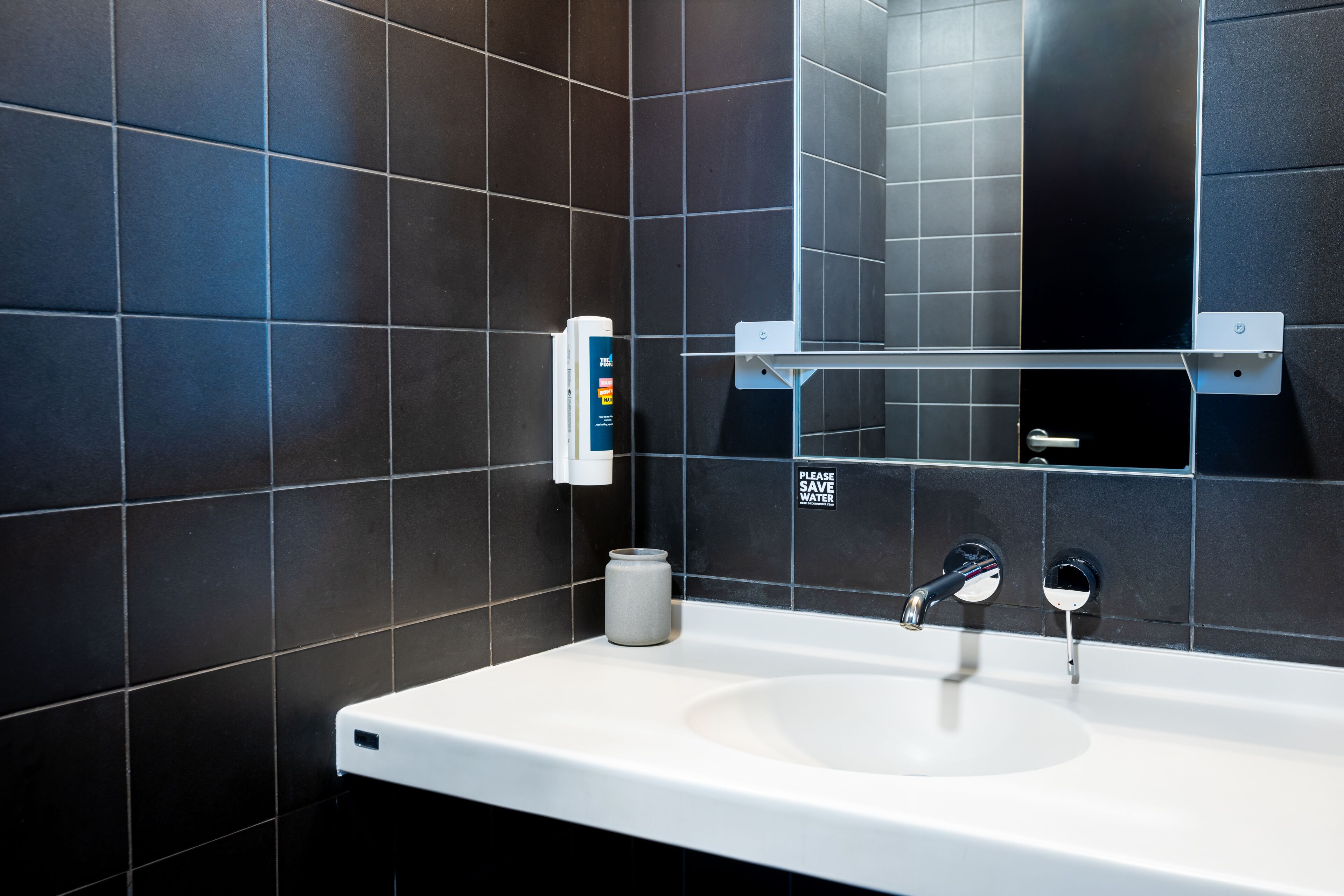 A modern bathroom with a white sink, dark tiles, a mirror, and a soap dispenser.