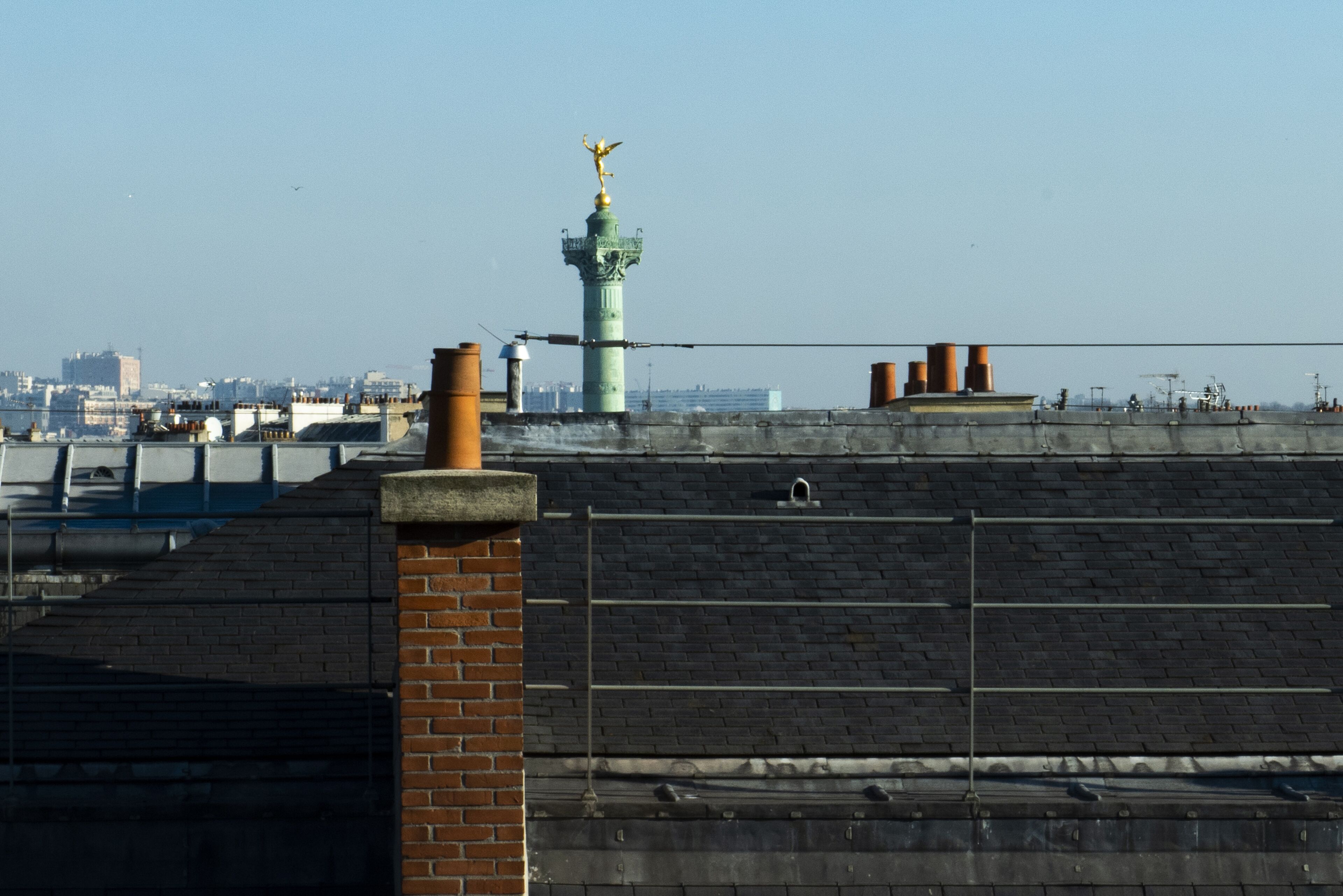 A rooftop with a brick chimney, a metal railing, and a green tower with a golden figure.