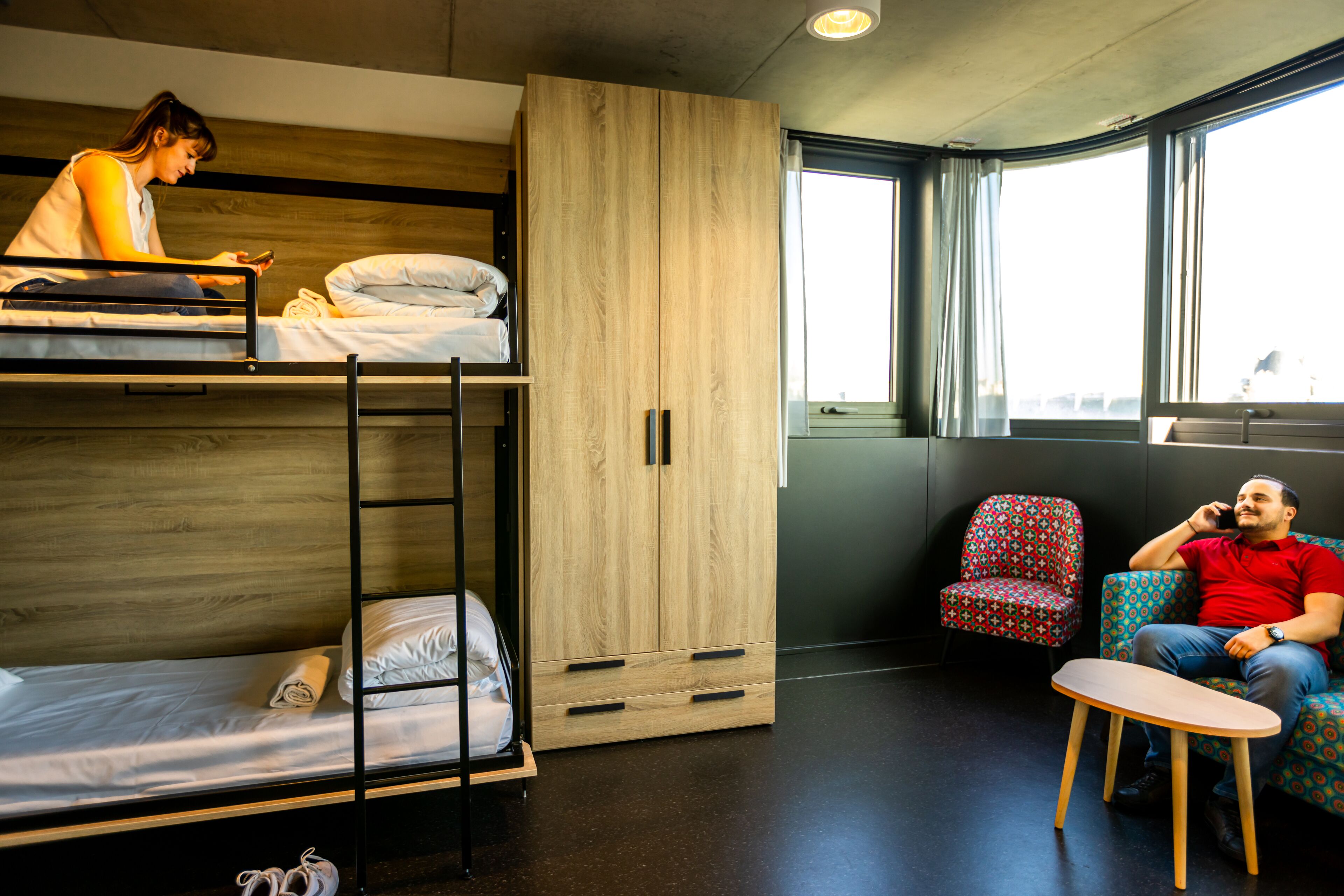 A bunk bed with a wooden wardrobe, a small table, and a patterned chair in a room with large windows.
