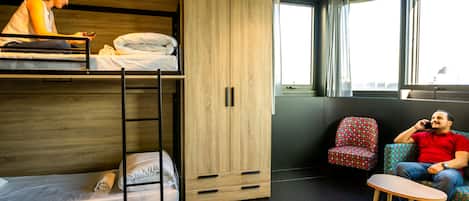 Cama tipo litera con un armario de madera, una mesita pequeña y una silla con estampado en una habitación con ventanas grandes.