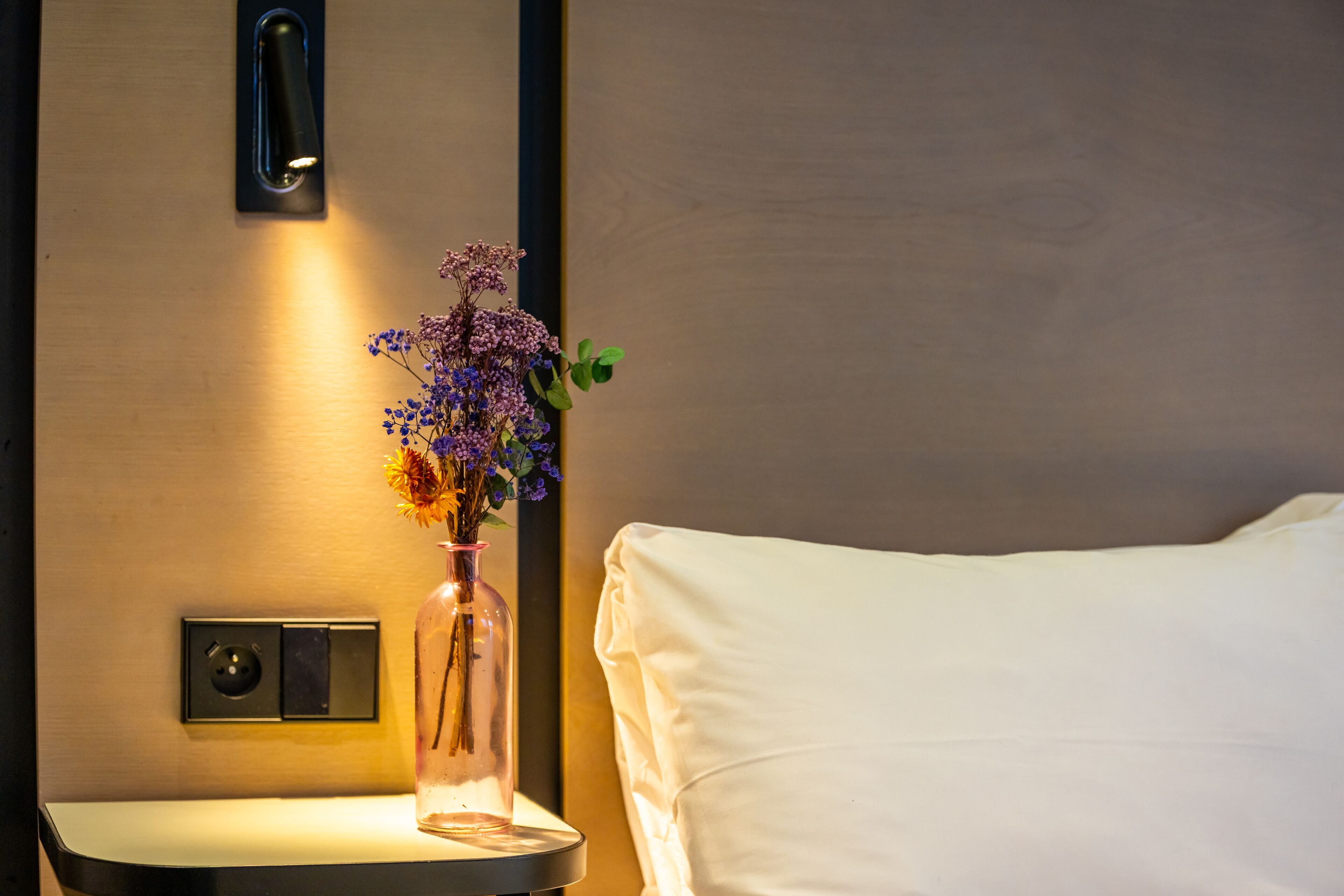 Habitación de hotel con mesita de noche, un jarrón con flores, una cama con sábanas blancas y un interruptor de luz en la pared.