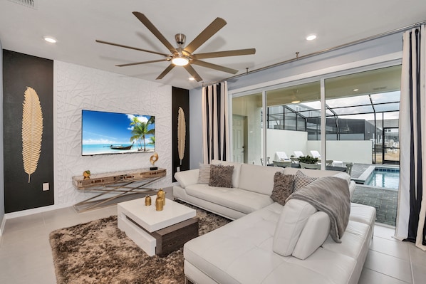 A modern living room with a white sofa, a ceiling fan, a flat-screen TV, and a view of a swimming pool.