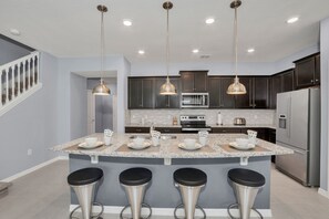 Una cucina moderna con piani in granito, elettrodomestici in acciaio inossidabile, illuminazione a sospensione e un'isola centrale con sgabelli da bar.