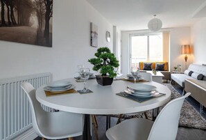 A dining area with a round table set for four, surrounded by white chairs. There is a small potted plant on the table, and the room has a white color scheme with a large window providing natural light.