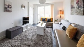 Ein modernes Wohnzimmer mit Flachbildfernseher, einer Couch, einem Couchtisch und einem großen Fenster mit Vorhängen.
