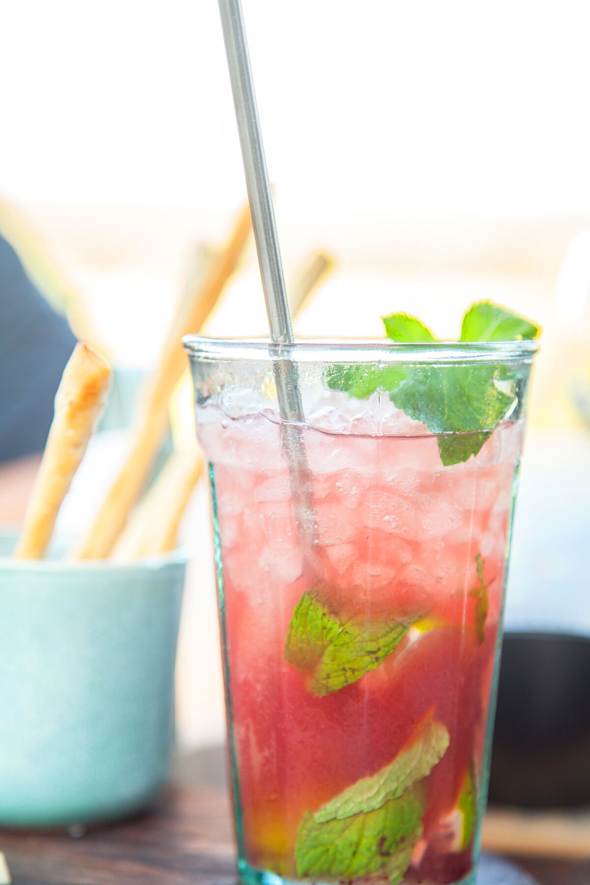 Ein Glas rosa Getränk mit Eiswürfeln und Minzblättern, dazu ein Metallstrohhalm und Baguettes in einer kleinen blauen Schüssel.