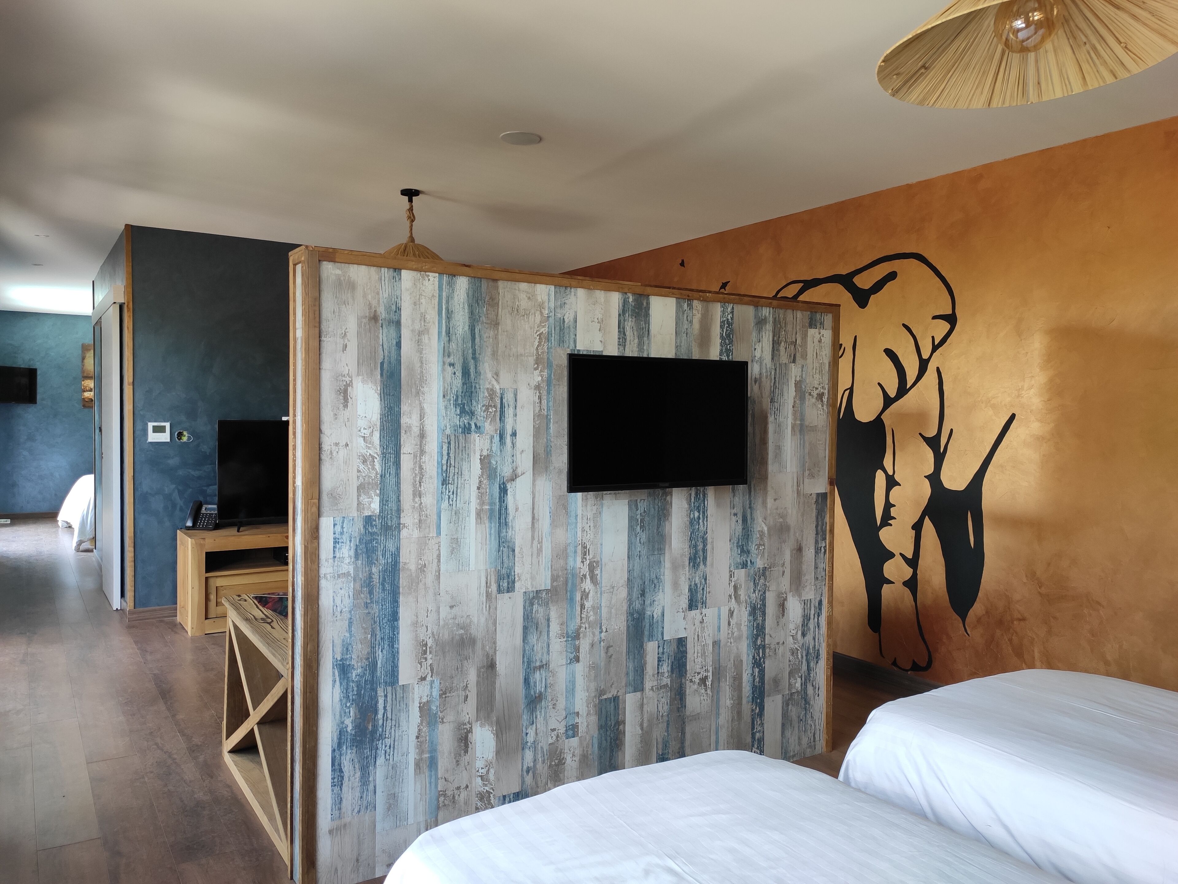 A hotel room with a bed, a TV mounted on a wooden panel wall, a ceiling fan, and a wall mural.