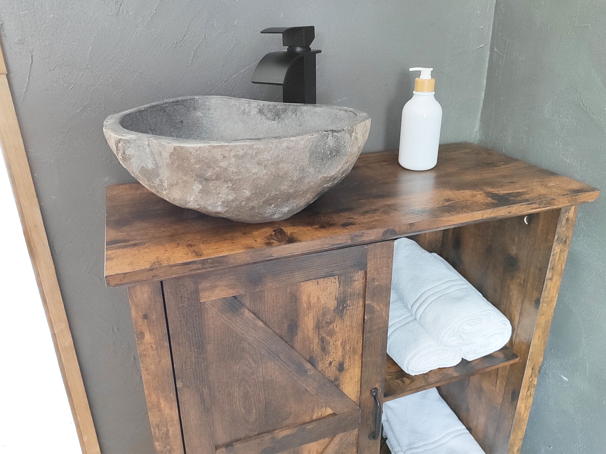 A wooden vanity with a large stone sink, a black faucet, and a white bottle of lotion.