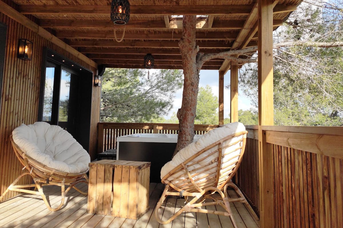 Une terrasse en bois avec deux fauteuils en osier et une petite table, un toit de chaume et une vue sur les arbres.