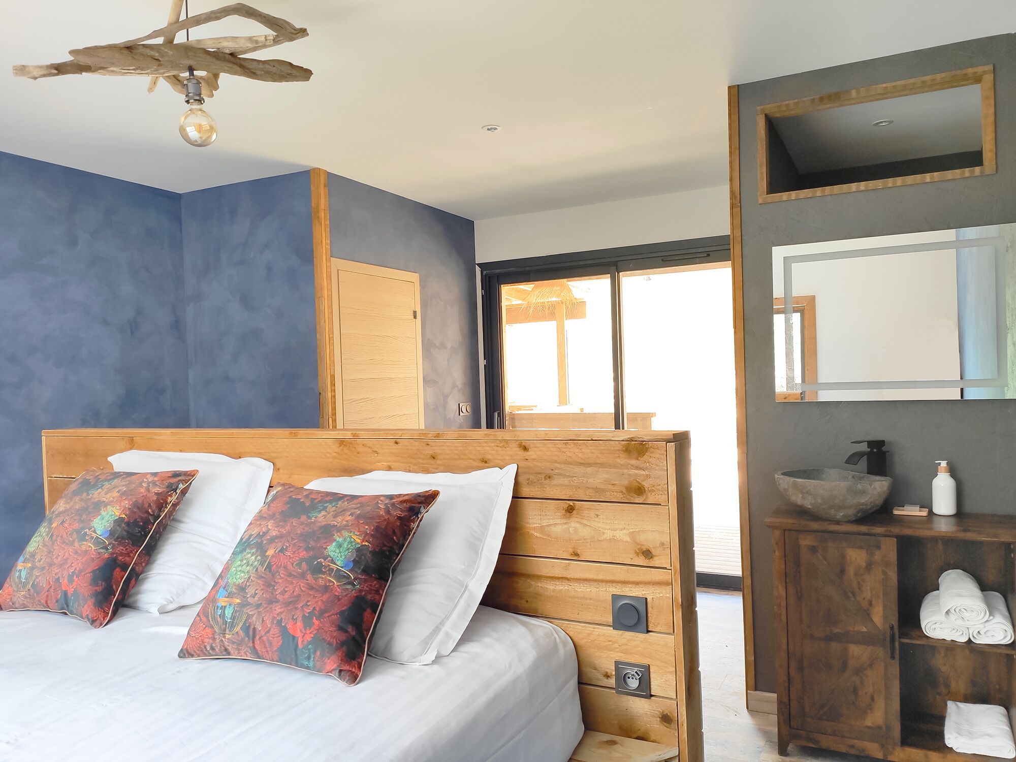 A bedroom with a wooden headboard, a large bed with white linens, two decorative pillows, a wooden nightstand with a bowl and toiletries, and a large mirror above the nightstand.