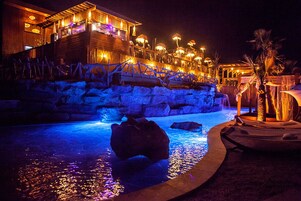 Vista noturna de um resort com piscina, deck de madeira e um edifício com janelas iluminadas.