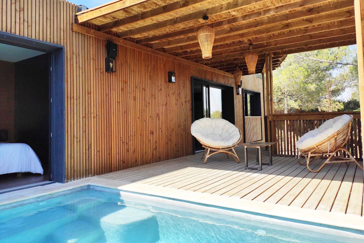 Une terrasse en bois avec une piscine, deux fauteuils en osier et un lit.