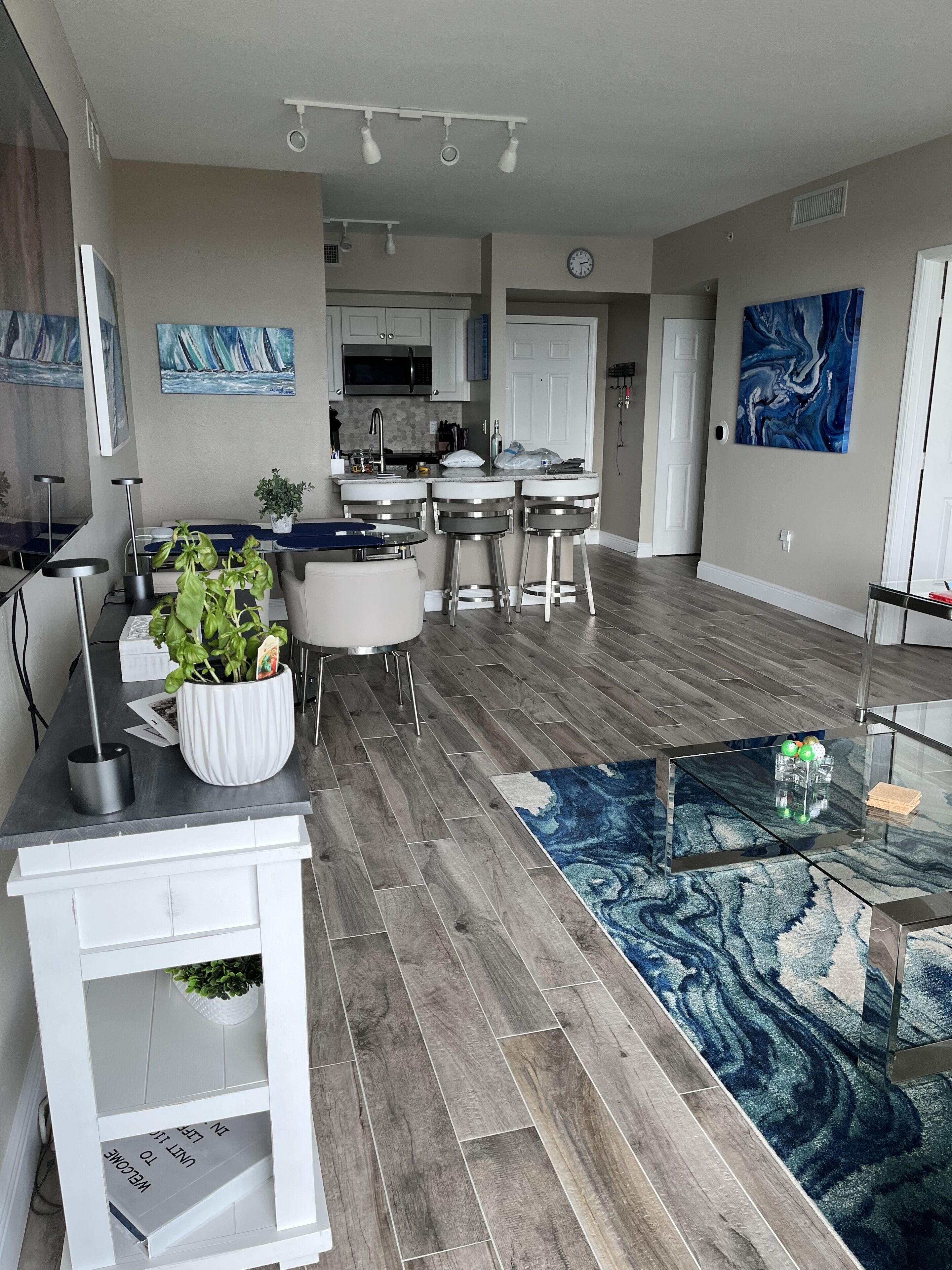 A modern kitchen with white cabinets, a microwave, and a sink. There is a dining area with a table and chairs, and a living room with a blue rug and a glass coffee table.