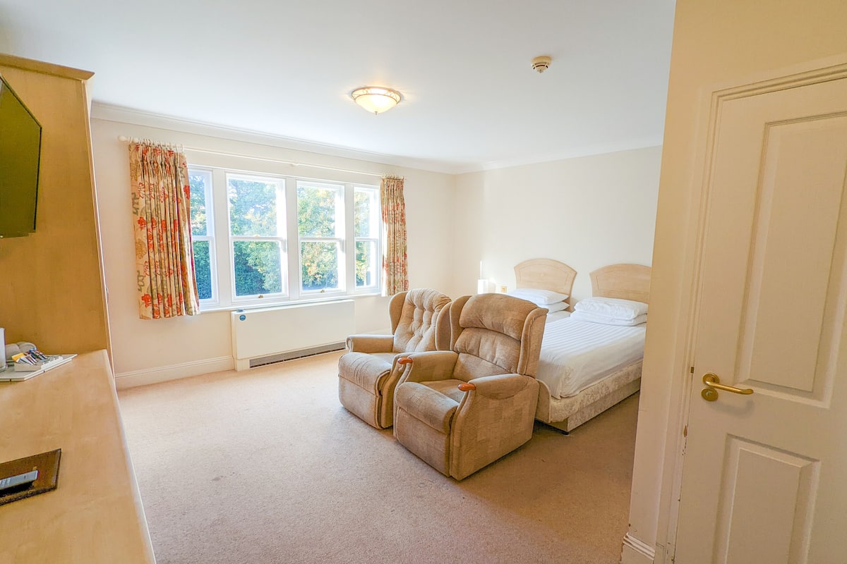 A bedroom with a bed, a chair, a radiator, a window with curtains, and a door.