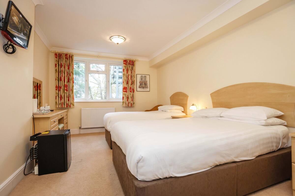 A hotel room with two beds, a desk, a radiator, and a window with curtains.