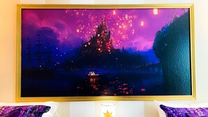 A hotel room with a large painting of a castle and lanterns, a bedside table with a lamp, and purple patterned pillows.