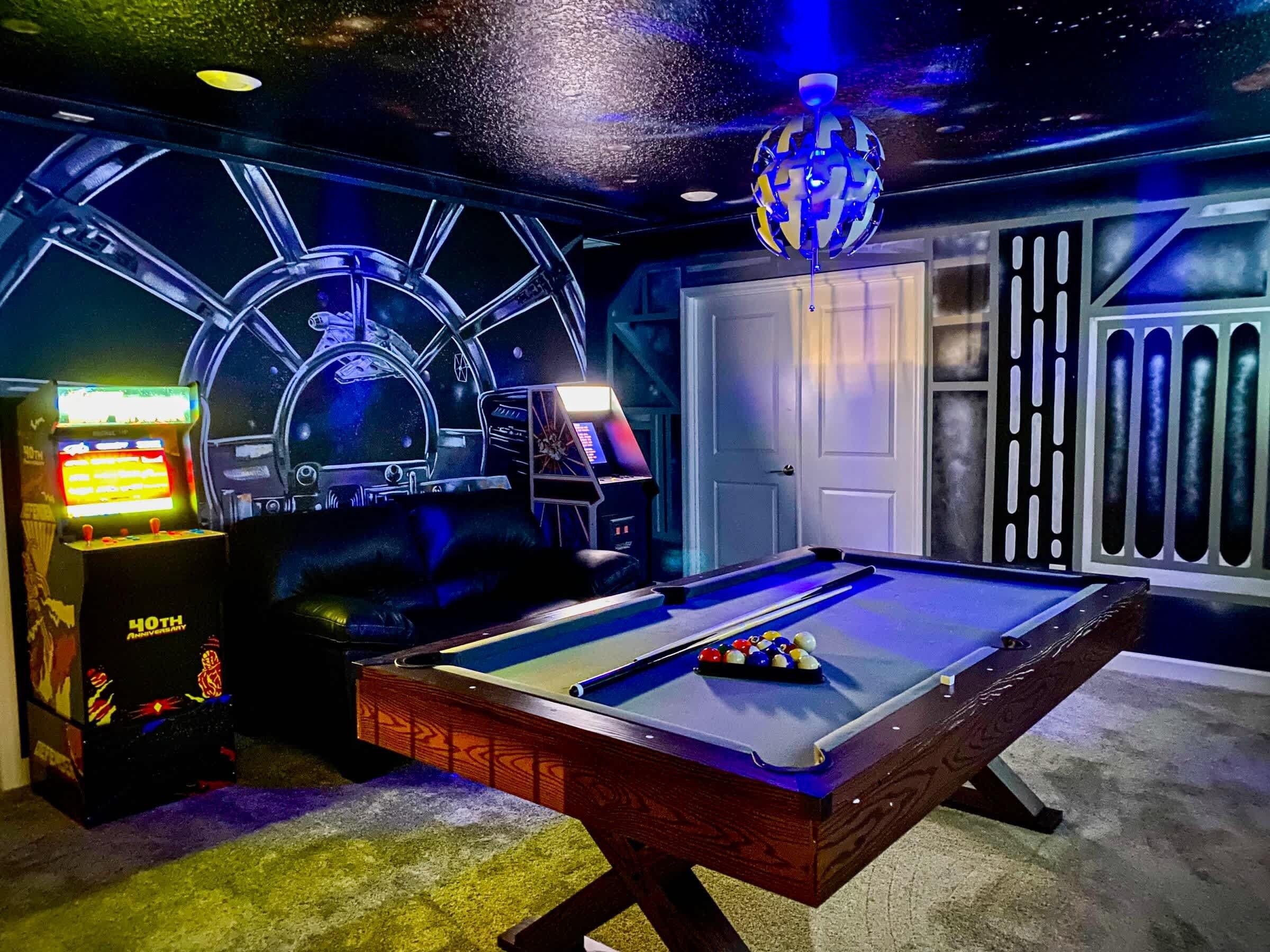 A pool table with a space-themed mural, a pool cue, and a video game machine.