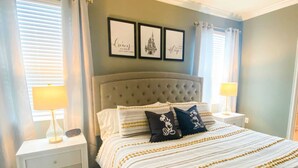 A bedroom with a tufted headboard, a bed with striped bedding, two bedside tables with lamps, and three framed pictures on the wall.